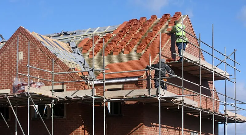A new brick house with its roof being tiled from scaffolding